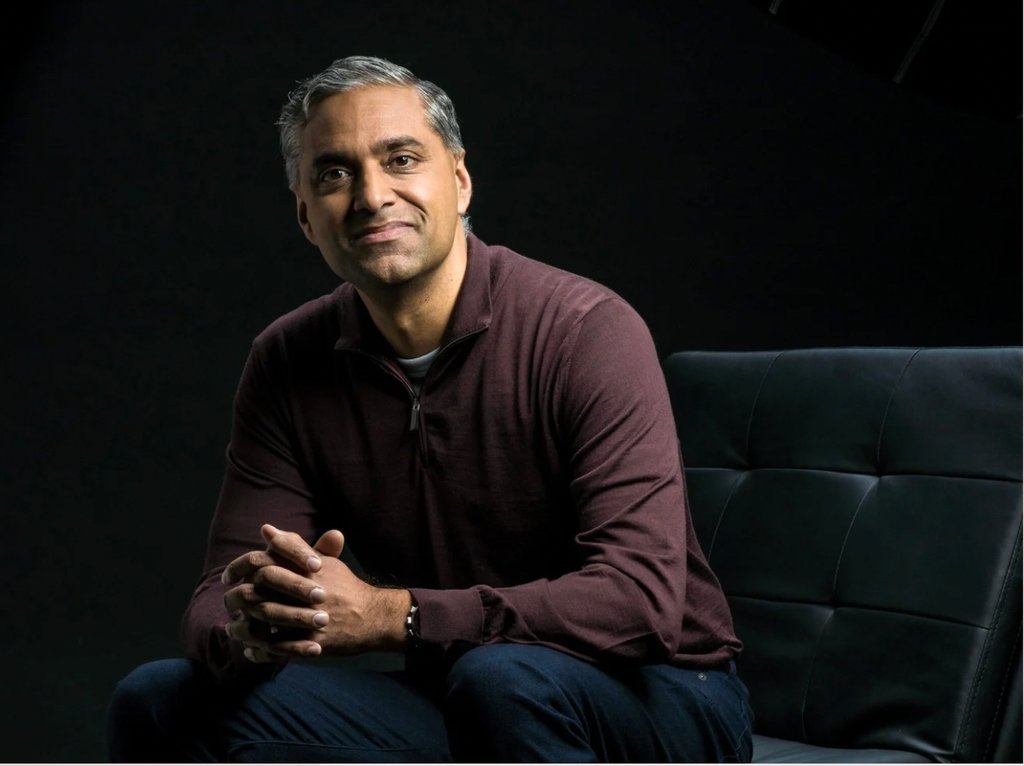 A man with short, gray hair sits on a black sofa, wearing a maroon zip-up sweater and dark pants, hands clasped and a slight smile—showcasing the confidence that comes from using the best personal styling service, set against a dark background.